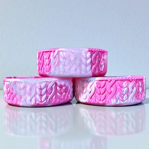May include: Three stacked, round containers with a pink and white marbled design. The containers have a textured, raised pattern resembling a knitted design. The containers are stacked on a reflective surface.