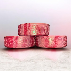 May include: Three stacked, oval-shaped containers in shades of pink and red. Each container has a floral pattern and a textured surface. The containers are arranged on a light-colored surface against a soft, gradient background.