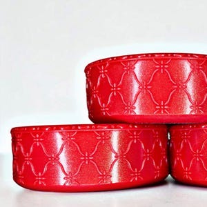 May include: Three stacked, bright red, round containers. Each container has a repeating floral pattern embossed on the sides. The containers are made of a solid material and have a slightly textured surface. The background is plain white.
