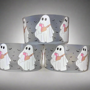 May include: Set of three frosted glass votive candle holders. Each holder features a white ghost illustration with a pink drink and a brown satchel. The background is a light gray with black bat silhouettes.