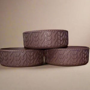 May include: Three stacked, round, brown containers with a leaf pattern. The containers are made of a matte material and have a textured surface. The containers are arranged in a triangular formation.