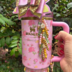 May include: Pink Stanley tumbler with floral design and the text "LoveShackFan". The tumbler is adorned with a pink bow with gold glitter accents, and beaded charms. The handle and other accessories are pink.