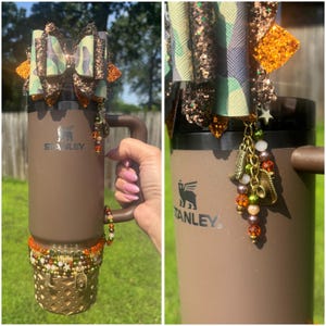 May include: A brown Stanley tumbler with a handle, adorned with a camouflage bow, glitter accents, and a charm cluster. The bottom features a decorative gold-colored wrap with beads. The tumbler is held by a person.