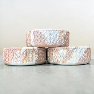 May include: Three round, shallow containers featuring a raised, textured cable knit pattern on the exterior. The surface has a marbled, pearlescent finish with swirling tones of soft peach, cream, and white, creating a subtle iridescent sheen.