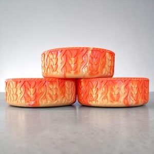 May include: Three round, shallow bowls with a vibrant orange and cream marbled finish. Each bowl features a raised, textured pattern of vertical wheat-like stalks or braided leaves encircling the exterior side walls in a repeating design.