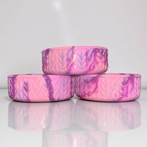 May include: Three round, decorative containers stacked. Each container is a blend of pink and purple hues, with a textured, woven pattern. The containers are arranged in a pyramid shape, with their reflections visible on the white surface.