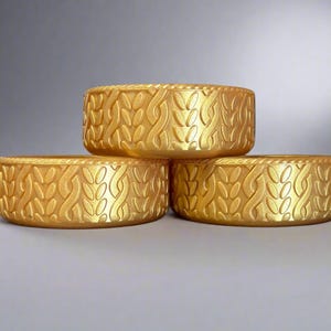May include: Three gold-colored, round decorative bowls. Each bowl has a repeating pattern of stylized leaves and vines. The bowls are stacked, with two on the bottom and one on top, creating a visually appealing arrangement.