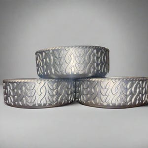 May include: Three silver-colored, round bowls with a repeating leaf-like pattern. The bowls are stacked, with two on the bottom and one on top. The bowls have a textured surface and a decorative edge.