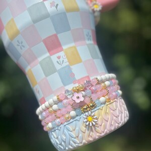 May include: A pastel-colored tumbler with a pink handle and floral accents. The tumbler features a checkered pattern in soft hues of pink, yellow, and blue. Adorning the base are beaded bracelets in shades of pink, white, and gold, with flower charms.