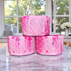 May include: Three pink decorative containers stacked on a marble surface. The containers have a textured design with diamond and wavy patterns in shades of pink and white. The background shows a window with a view of trees.