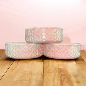 May include: Three decorative, round bowls stacked on a wooden surface. The bowls are white and pink with a textured, woven pattern. The background is a solid pink color.