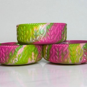 May include: Three small, oval-shaped containers in a stack. The containers are decorated with a textured, woven pattern and are colored with a blend of pink, green, and yellow. The containers are likely for storage or display.
