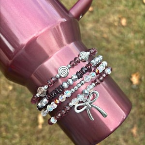 May include: A set of five beaded bracelets with silver accents. The bracelets are layered on a pink tumbler. The bracelets feature clear, iridescent, and dark purple beads, as well as silver swirl and bow charms.