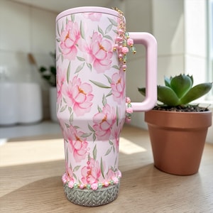 May include: A pink insulated tumbler with a floral peony design. The tumbler has a pink handle and lid, and is adorned with beaded charms. A small succulent in a terracotta pot sits nearby.