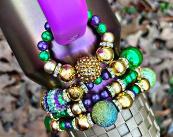 May include: A set of four beaded bracelets in green, purple, gold, and clear rhinestone colors. The bracelets are stacked on a purple and gold tumbler.