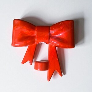 May include: A bright red, glossy bow-shaped object with a cylindrical piece below it. The bow has a classic design with loops and tails. The object is likely a decorative element or a part of a larger design.