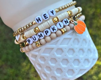 Może przedstawiać: Zestaw czterech bransoletek z złotymi koralikami i białymi koralikami, które tworzą napis "HEY PUMPKIN" oraz mały pomarańczowy charms w kształcie dyni.