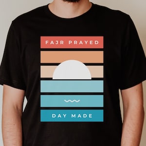May include: A black t-shirt with a graphic design featuring a sunset with a white sun and blue waves. The text "FAJR PRAYED" and "DAY MADE" is printed on the shirt.