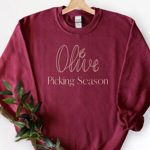 Op de afbeelding: Een bordeauxrode sweatshirt met de woorden "Olive Picking Season" in een gouden scriptlettertype. Het woord "Olive" heeft een kleine olijftak-afbeelding. De sweatshirt hangt aan een houten hanger.