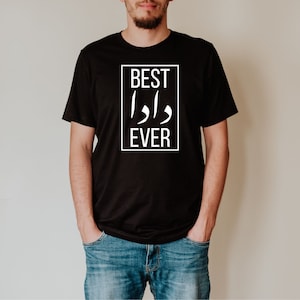 May include: Black t-shirt with the text "BEST DADA EVER" in a white rectangular frame. The shirt is a crew neck style and is being worn by a person. The person is wearing blue jeans.