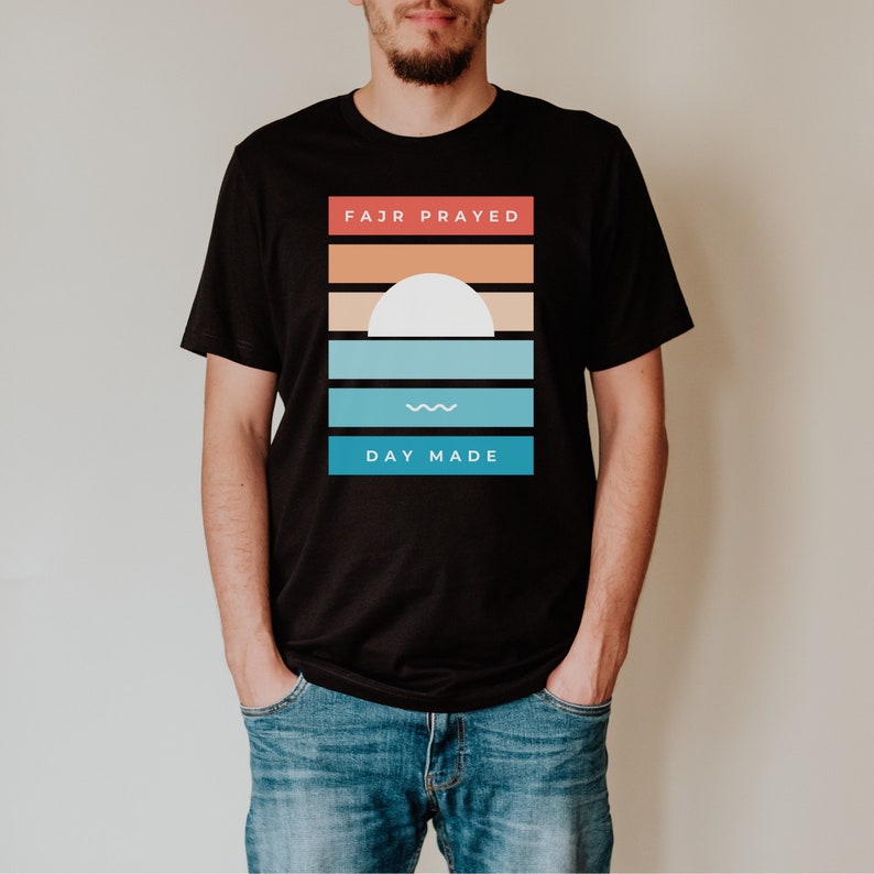 May include: A black t-shirt with a graphic design featuring a sunset with a white sun and blue waves. The text "FAJR PRAYED" and "DAY MADE" is printed on the shirt.