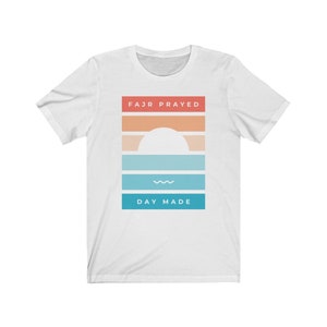 May include: White t-shirt with a graphic design featuring a sunset with a white sun and a wavy line. The text "FAJR PRAYED" is above the sunset and "DAY MADE" is below the sunset.