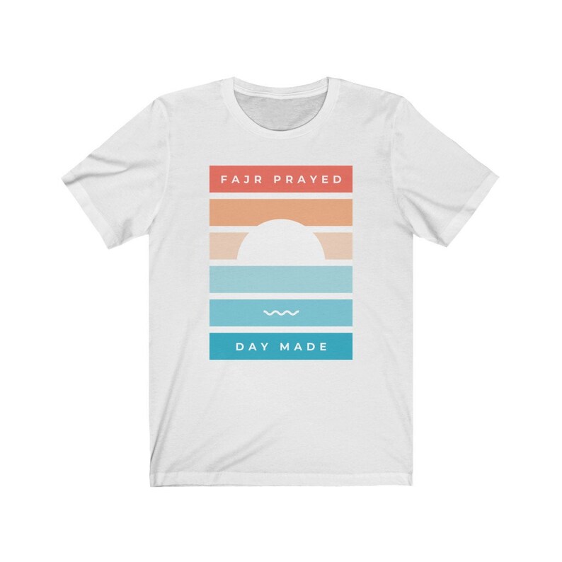 May include: White t-shirt with a graphic design featuring a sunset with a white sun and a wavy line. The text "FAJR PRAYED" is above the sunset and "DAY MADE" is below the sunset.