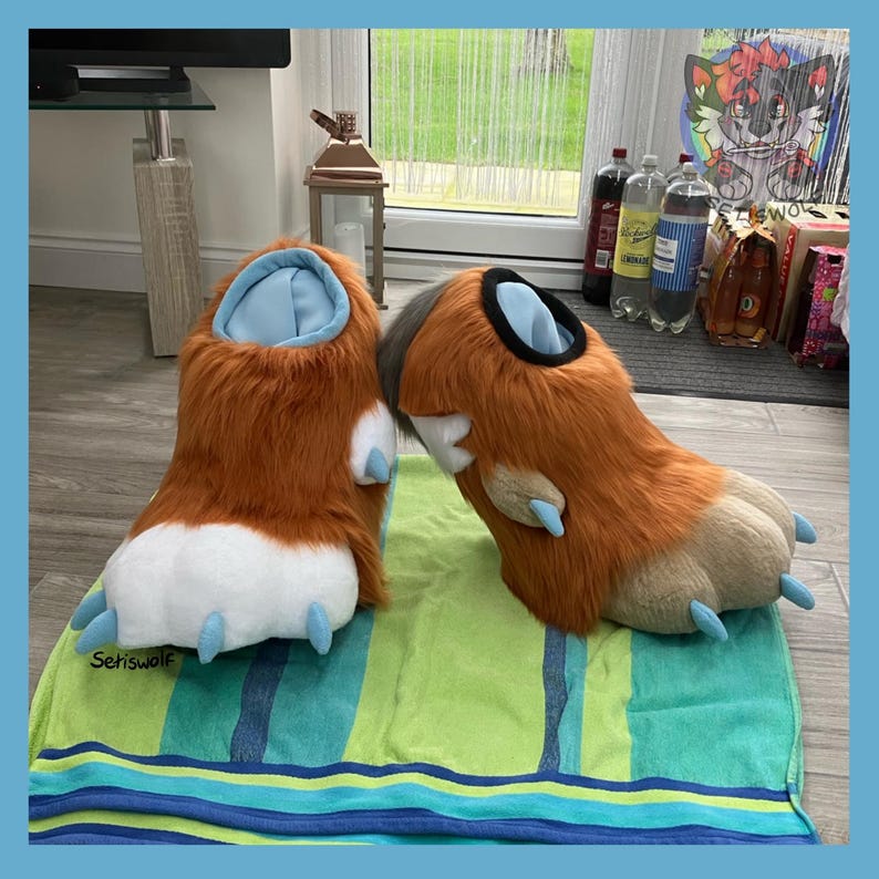 May include: A pair of furry, brown and tan paw-shaped slippers with light blue accents. The slippers have white pads and light blue claws. The slippers are resting on a green and blue striped towel. The text "Setiswolf" is visible on the towel.