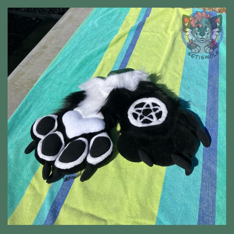 May include: A pair of black and white faux fur animal paw gloves. One glove features a white heart-shaped palm pad with black paw pads, while the other displays a white pentagram symbol on a black circular patch. Both gloves have black claws.