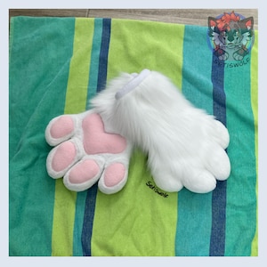 May include: A pair of white faux fur animal paw gloves featuring soft pink padded paw pads and a heart-shaped palm pad. The gloves have a thick, plush texture and a white cuff. The text Setiswolf is printed in black on the lower right side.