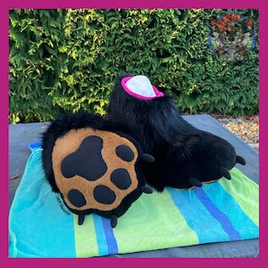 May include: Black furry bear paw costume feet with a brown and black paw pad. The feet have a pink and white interior and are displayed on a striped blue and green towel. The costume is designed for cosplay or costume parties.