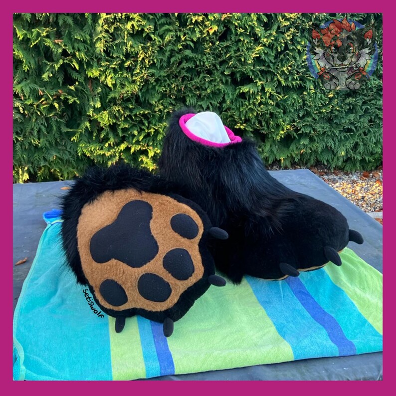 May include: Black furry bear paw costume feet with a brown and black paw pad. The feet have a pink and white interior and are displayed on a striped blue and green towel. The costume is designed for cosplay or costume parties.