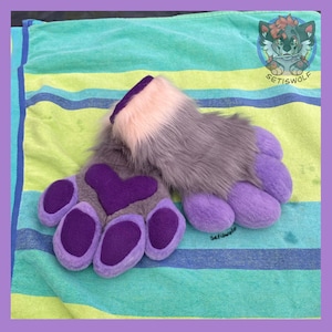 May include: A pair of plush faux fur paw gloves featuring grey fur, a light pink cuff, and purple paw pads with a heart-shaped palm pad. The design includes a small circular logo in the corner with a wolf illustration and the text SETISWOLF.