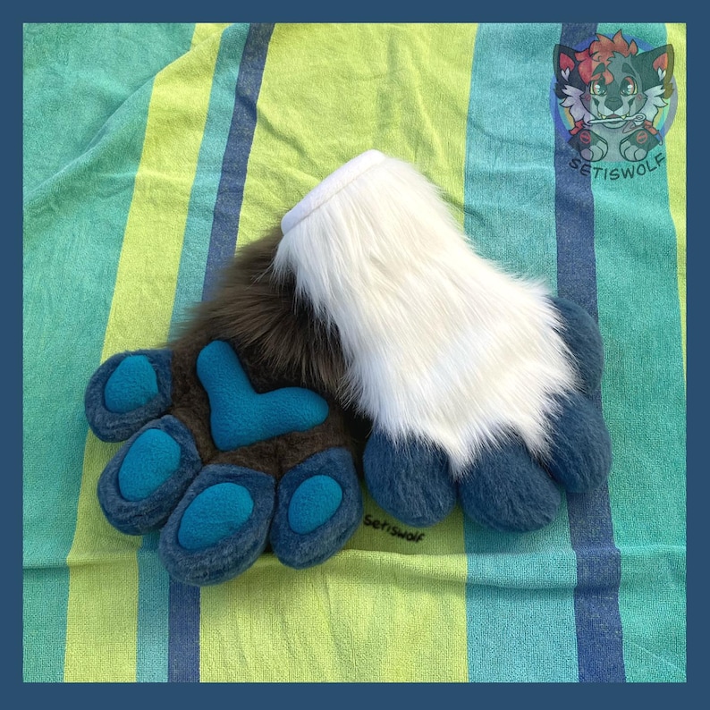 May include: A pair of plush animal paw gloves featuring long white faux fur on the upper section and dark brown faux fur on the lower section. The palm area is dark brown with bright teal blue padded paw pads and a central heart-shaped pad.