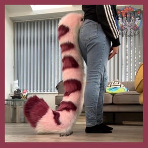 May include: A large, plush tail in shades of pink and burgundy, designed with alternating stripes. The tail is fluffy and appears to be made of faux fur. The tail is being worn by a person wearing blue jeans and a black top.