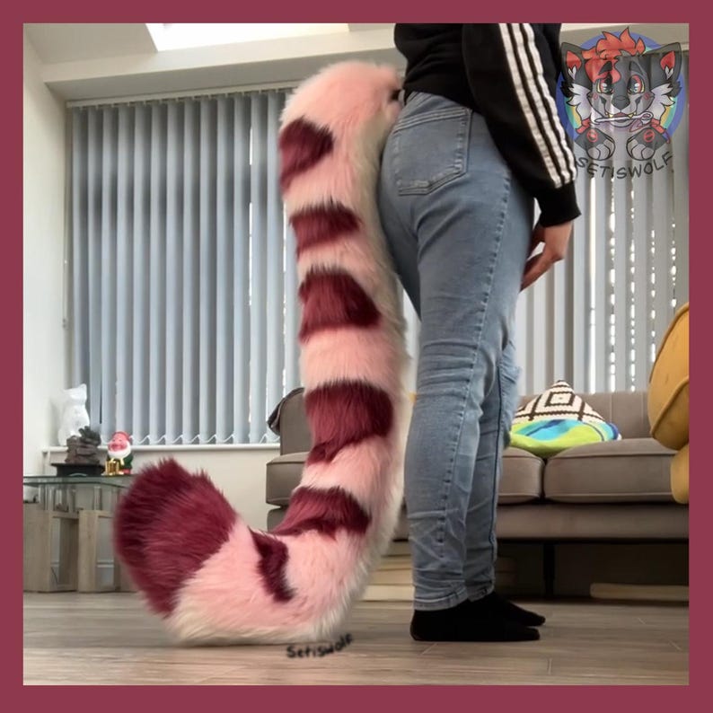 May include: A large, plush tail in shades of pink and burgundy, designed with alternating stripes. The tail is fluffy and appears to be made of faux fur. The tail is being worn by a person wearing blue jeans and a black top.