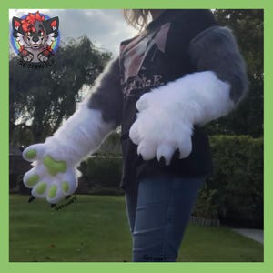May include: A pair of long, plush, grey and white faux fur animal hand paws featuring bright lime green paw pads and black claws. The sleeves extend up the forearm, and the paws are worn with a black t-shirt and blue denim jeans.