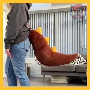 May include: A brown and yellow plush tail, likely for a costume, is attached to a person's waist. The tail is a reddish-brown color with yellow accents. The tail is long and curved, with a fluffy texture. The person is wearing a gray hoodie and blue jeans.