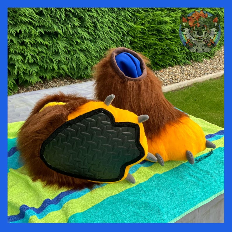 May include: A pair of furry brown and orange paw-shaped costume feet. The paws have yellow and black details, with gray claws and a textured black sole. The interior is blue. The feet are resting on a striped green and blue towel.