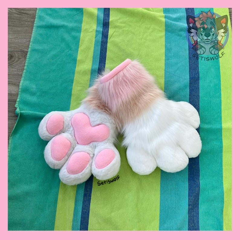 May include: A pair of plush animal paw gloves featuring white faux fur, a soft pink faux fur cuff, and pink padded paw pads. The design includes four rounded fingers and a central heart-shaped palm pad, with the text Setiswolf visible.