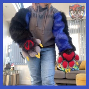 May include: A pair of long, plush faux fur sleeves featuring a color-blocked pattern of black and vibrant blue. The attached paw-shaped gloves have grey fur, red paw pads, and yellow claws. A grey hooded sweatshirt and blue jeans are worn.