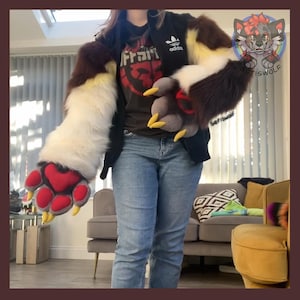 May include: A pair of long, plush faux fur sleeves featuring a brown, white, and yellow color scheme with large, padded grey paw gloves. The paws have red heart-shaped pads and sharp yellow claws. The sleeves are worn over a black jacket and jeans.