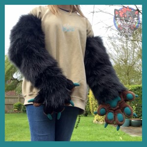 May include: A pair of long black faux fur arm sleeves with attached paw-shaped gloves. The gloves feature brown fabric palms, teal paw pads, and matching teal claws. The sleeves extend from the wrists up to the upper arms.