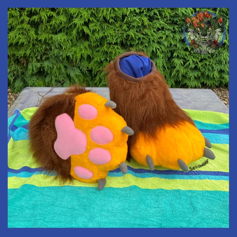 May include: A pair of furry, brown and yellow paw-shaped costume feet. The paws have pink pads and gray claws. The top of the feet are brown with a blue interior. The paws are on a striped towel.