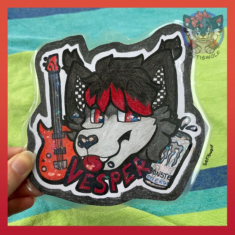K&ouml;nnte beinhalten: Ein transparenter Acryl-Anh&auml;nger mit einem schwarz-grauen anthropomorphen Wolfscharakter mit roten Haarstr&auml;hnen, einer roten E-Gitarre und einer Energy-Drink-Dose. Der Name VESPER ist in Rot unter dem Charakter aufgedruckt, mit glitzerndem Finish.