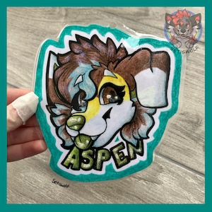 K&ouml;nnte beinhalten: Ein durchsichtiger Acryl-Badge mit einer handgezeichneten Illustration eines pelzigen Charakters mit braunem, wei&szlig;em und gelbem Fell, gro&szlig;en dunklen Augen und einer gr&uuml;nen Nase. Der Name ASPEN ist in fetten gr&uuml;nen Buchstaben aufgedruckt.
