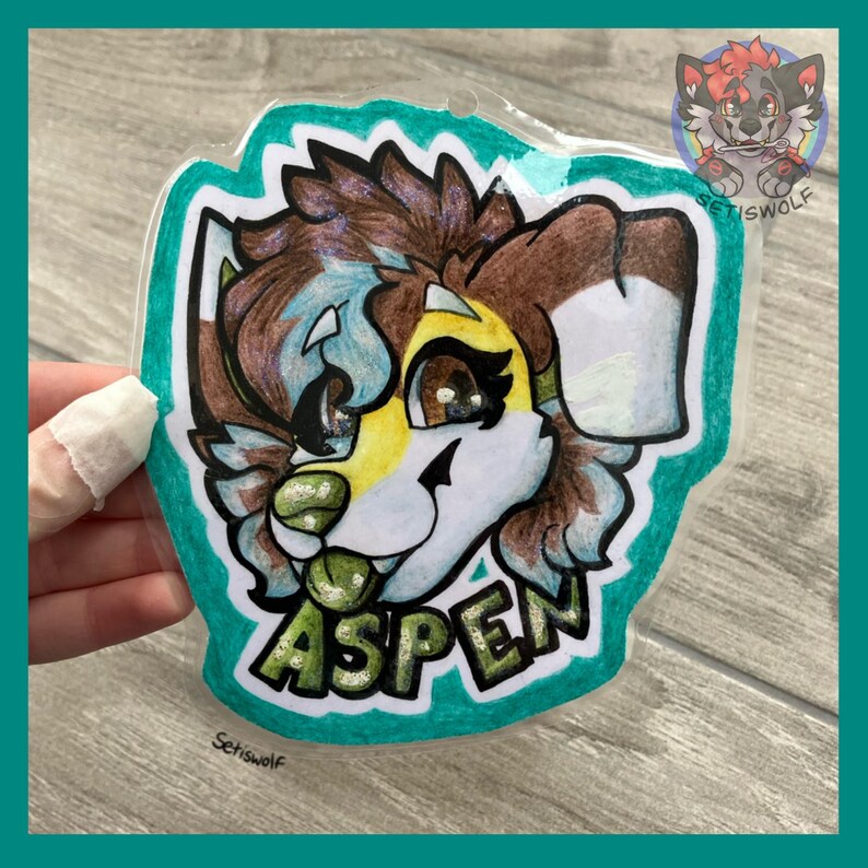 K&ouml;nnte beinhalten: Ein durchsichtiger Acryl-Badge mit einer handgezeichneten Illustration eines pelzigen Charakters mit braunem, wei&szlig;em und gelbem Fell, gro&szlig;en dunklen Augen und einer gr&uuml;nen Nase. Der Name ASPEN ist in fetten gr&uuml;nen Buchstaben aufgedruckt.