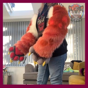 May include: A pair of long, plush, orange and white faux fur sleeves with attached paw-shaped gloves. The gloves feature red and grey padded paw pads and pointed yellow claws. The sleeves are worn over a black hooded sweatshirt and blue jeans.