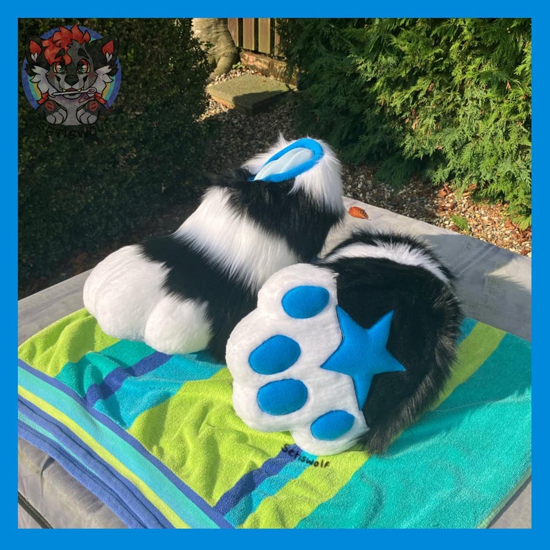 May include: A pair of black and white furry paw pads with blue accents. The paw pads have a star and circular details in blue. The items are placed on a striped green and blue towel.
