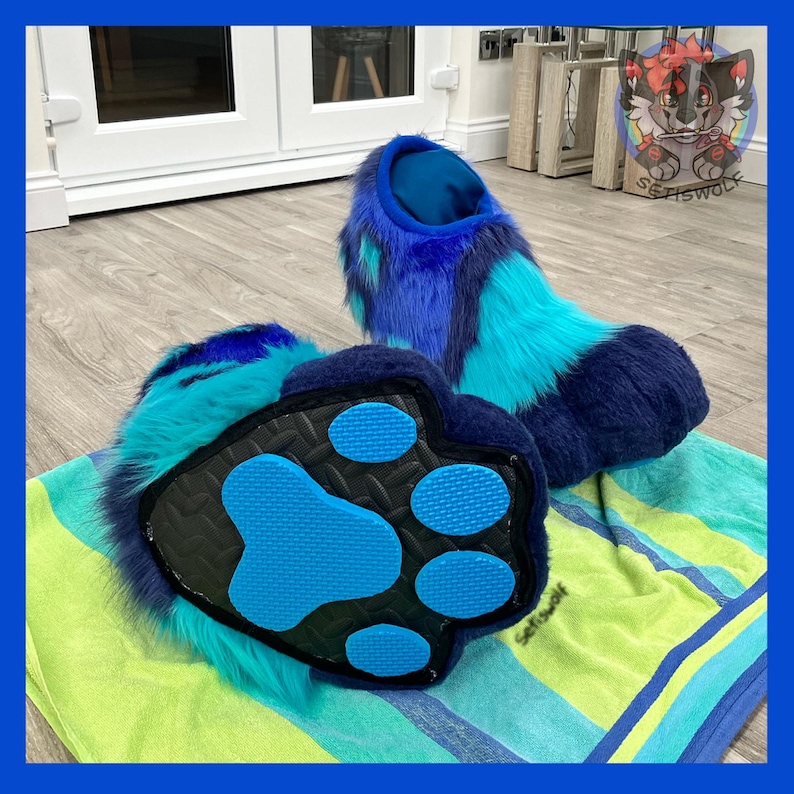 May include: A pair of blue and teal furry paw-shaped slippers. The slippers have a dark blue paw pad with light blue paw prints. The fur is a mix of dark blue, teal, and light blue. The slippers are designed to resemble animal paws.